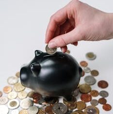Hand inserting coin into a black piggy bank surrounded by various coins, symbolizing savings.